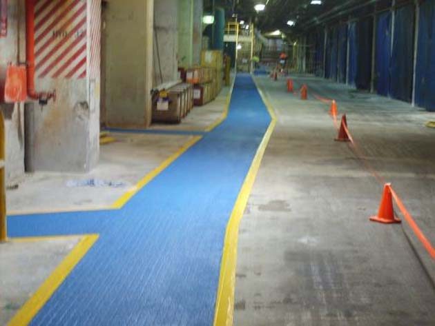 A blue anti-skid pedestrian walkway with yellow edge lines through an industrial plant, orange cones alongside