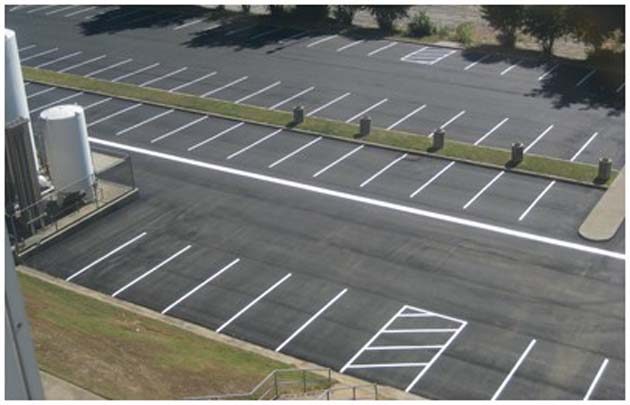 A car park seen from above with white bay lines and a hatched area beside a building and storage tanks