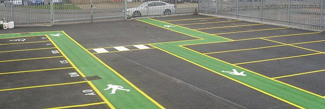 Green painted pedestrian walkways with white walking symbols and yellow bay lines in a fenced car park