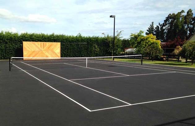 A dark asphalt tennis court with white lines, a net and a timber practice wall against a hedge
