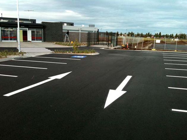 Two white directional arrows and parking bay lines on new black asphalt outside a modern commercial building